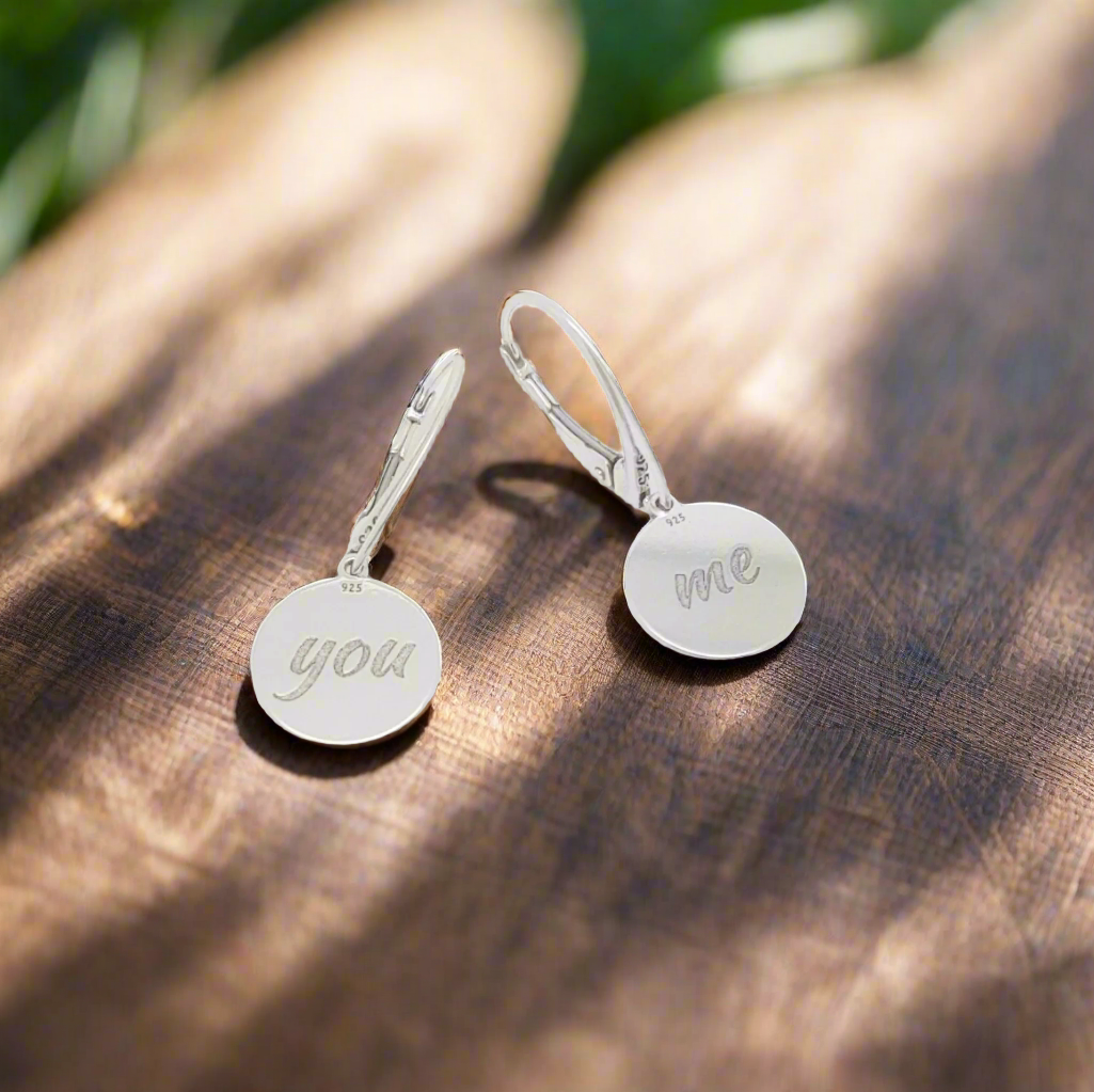 Close-up of You and Me Sterling Silver Drop Earrings highlighting engraved text "you" and "me" in minimalist style. "You & Me" engraved sterling silver drop earrings with leverback design, displayed on a wooden surface